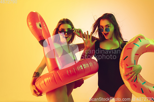 Image of Beautiful girls in fashionable swimsuits isolated on yellow studio background in neon light. Summer, resort, fashion and weekend concept. Posing in rubber donut and flamingo with bright emotions.