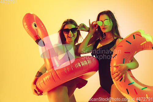 Image of Beautiful girls in fashionable swimsuits isolated on yellow studio background in neon light. Summer, resort, fashion and weekend concept. Posing in rubber donut and flamingo with bright emotions.