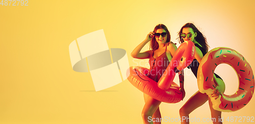 Image of Beautiful girls in fashionable swimsuits isolated on yellow studio background in neon light. Summer, resort, fashion and weekend concept. Posing in rubber donut and flamingo, flyer with copyspace.