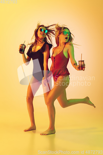 Image of Beautiful girls in fashionable swimsuits isolated on yellow studio background in neon light. Summer, resort, fashion and weekend concept. With cocktails on the run.