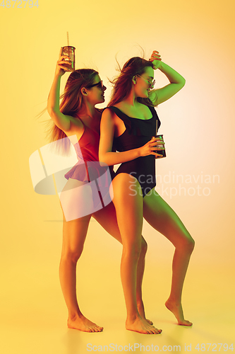 Image of Beautiful girls in fashionable swimsuits isolated on yellow studio background in neon light. Summer, resort, fashion and weekend concept. Dancing with cocktails.