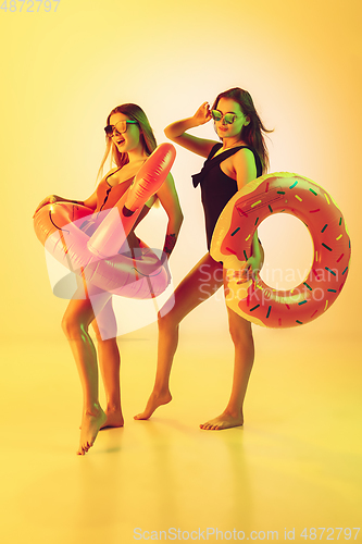 Image of Beautiful girls in fashionable swimsuits isolated on yellow studio background in neon light. Summer, resort, fashion and weekend concept. Posing in rubber donut and flamingo with bright emotions.