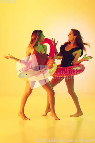 Image of Beautiful girls in fashionable swimsuits isolated on yellow studio background in neon light. Summer, resort, fashion and weekend concept. Posing in rubber donut and flamingo with bright emotions.