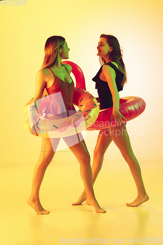 Image of Beautiful girls in fashionable swimsuits isolated on yellow studio background in neon light. Summer, resort, fashion and weekend concept. Posing in rubber donut and flamingo with bright emotions.