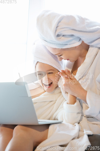 Image of Mother and daughter, sisters have quite, beauty and fun day together at home. Comfort and togetherness. Watching series using laptop near window wearing white bathrobes