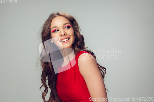 Image of Beautiful young woman with long healthy curly hair and bright make up wearing red dress isolated on grey studio backgroud. Smiling.