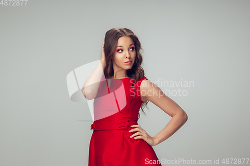 Image of Beautiful young woman with long healthy curly hair and bright make up wearing red dress isolated on grey studio backgroud. Calm, thoughtful.