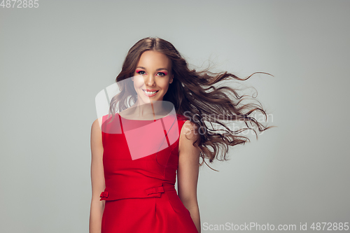 Image of Beautiful young woman with long healthy curly hair and bright make up wearing red dress isolated on grey studio backgroud. Flying, blowing hair.