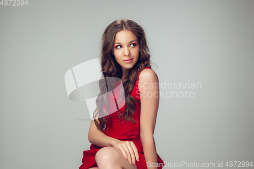 Image of Beautiful young woman with long healthy curly hair and bright make up wearing red dress isolated on grey studio backgroud. Calm, thoughtful.