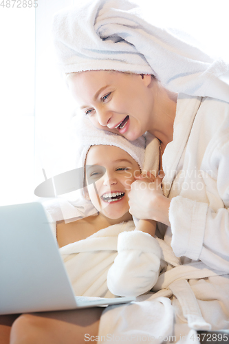 Image of Mother and daughter, sisters have quite, beauty and fun day together at home. Comfort and togetherness. Watching series using laptop near window wearing white bathrobes