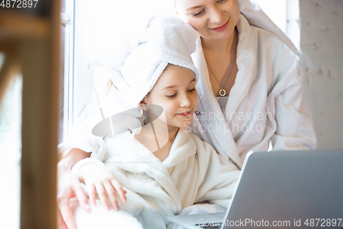 Image of Mother and daughter, sisters have quite, beauty and fun day together at home. Comfort and togetherness. Watching series using laptop near window wearing white bathrobes