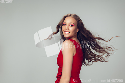 Image of Beautiful young woman with long healthy curly hair and bright make up wearing red dress isolated on grey studio backgroud. Flying, blowing hair.