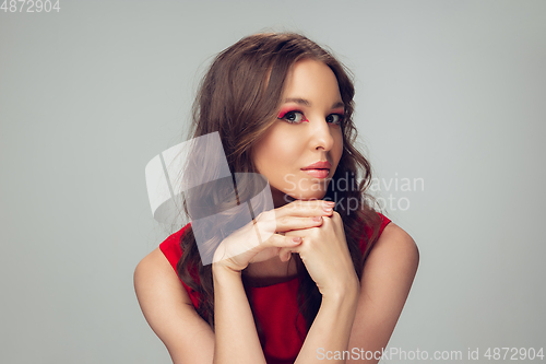 Image of Beautiful young woman with long healthy curly hair and bright make up wearing red dress isolated on grey studio backgroud. Calm, thoughtful.