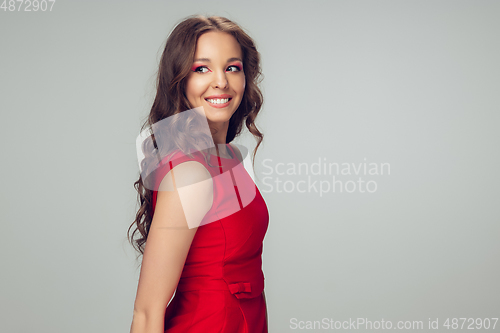 Image of Beautiful young woman with long healthy curly hair and bright make up wearing red dress isolated on grey studio backgroud. Smiling.