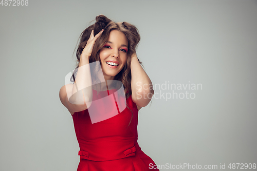 Image of Beautiful young woman with long healthy curly hair and bright make up wearing red dress isolated on grey studio backgroud. Smiling.