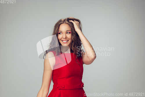 Image of Beautiful young woman with long healthy curly hair and bright make up wearing red dress isolated on grey studio backgroud. Smiling.