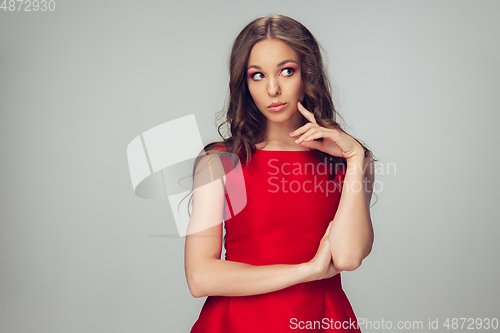 Image of Beautiful young woman with long healthy curly hair and bright make up wearing red dress isolated on grey studio backgroud. Calm, thoughtful.