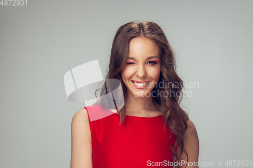 Image of Beautiful young woman with long healthy curly hair and bright make up wearing red dress isolated on grey studio backgroud, winking, smiling