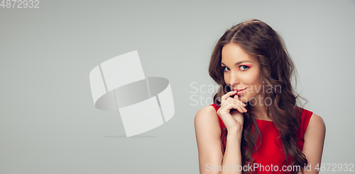 Image of Beautiful young woman with long healthy curly hair and bright make up wearing red dress isolated on grey studio backgroud. Flyer with copyspace.