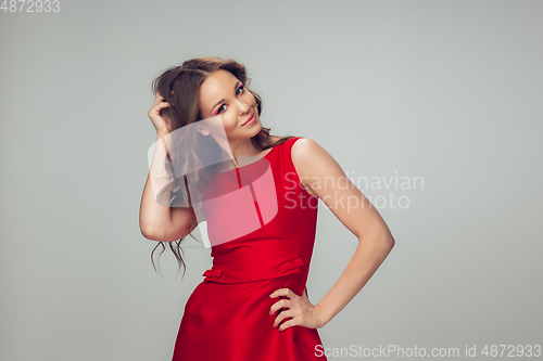 Image of Beautiful young woman with long healthy curly hair and bright make up wearing red dress isolated on grey studio backgroud. Shiny and confident.