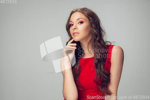 Image of Beautiful young woman with long healthy curly hair and bright make up wearing red dress isolated on grey studio backgroud. Calm, thoughtful.