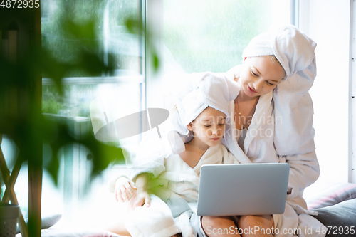 Image of Mother and daughter, sisters have quite, beauty and fun day together at home. Comfort and togetherness. Watching series using laptop near window wearing white bathrobes