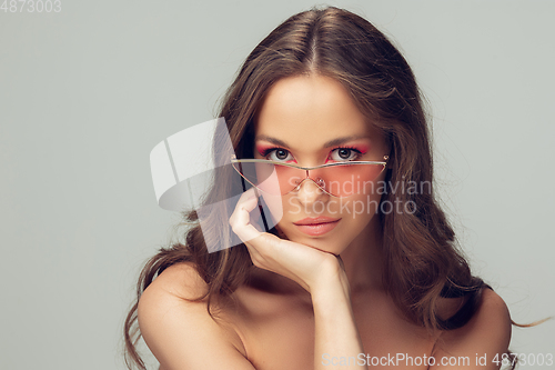 Image of Close up of beautiful young woman with long healthy curly hair and bright make up wearing stylish pink eyewear isolated on grey studio backgroud