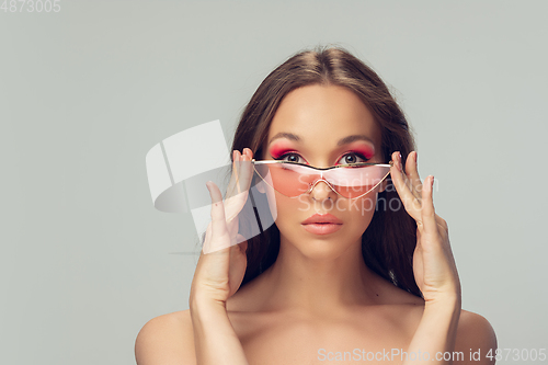 Image of Close up of beautiful young woman with long healthy curly hair and bright make up wearing stylish pink eyewear isolated on grey studio backgroud