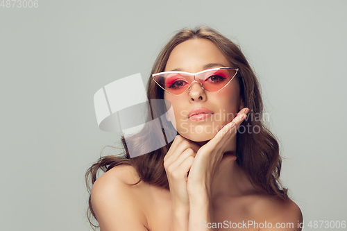 Image of Close up of beautiful young woman with long healthy curly hair and bright make up wearing stylish pink eyewear isolated on grey studio backgroud