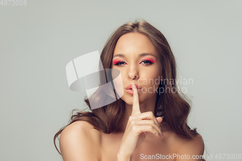 Image of Close up of beautiful young woman with long healthy curly hair and bright make up isolated on grey studio backgroud, whispering a secret with finger on lips