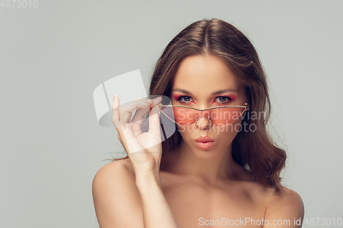 Image of Close up of beautiful young woman with long healthy curly hair and bright make up wearing stylish pink eyewear isolated on grey studio backgroud, blowing