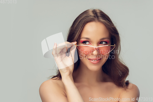Image of Close up of beautiful young woman with long healthy curly hair and bright make up wearing stylish pink eyewear isolated on grey studio backgroud, smiling