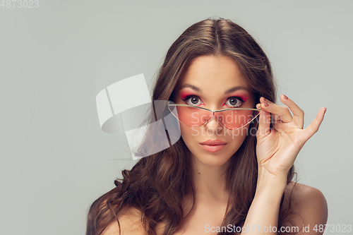 Image of Close up of beautiful young woman with long healthy curly hair and bright make up wearing stylish pink eyewear isolated on grey studio backgroud
