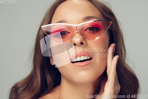 Image of Close up of beautiful young woman with long healthy curly hair and bright make up wearing stylish pink eyewear isolated on grey studio backgroud, smiling