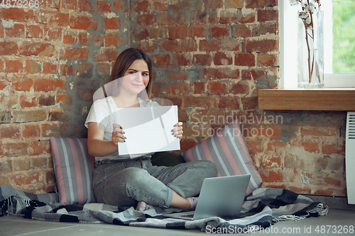 Image of Young woman, businesswoman working or studying at home, looking on computer screen, monitor, holding white sheet, whiteboard. Attented, concentrated. Copyspace. Top view.