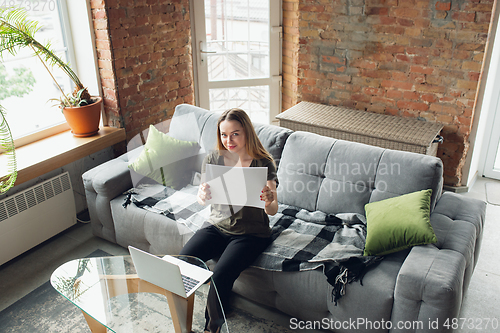 Image of Young woman, businesswoman working or studying at home, looking on computer screen, monitor, holding white sheet, whiteboard. Attented, concentrated. Copyspace. Top view.