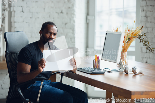 Image of Young man, businessman working in office, looking on blank black computer screen, monitor, holding blank white sheet, whiteboard. Copyspace.