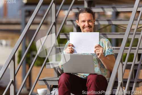 Image of Young man, businessman working outdoors, looking on computer screen, monitor, holding blank white sheet, whiteboard. Copyspace.