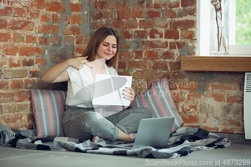 Image of Young woman, businesswoman working or studying at home, looking on computer screen, monitor, holding white sheet, whiteboard. Attented, concentrated. Copyspace. Top view.