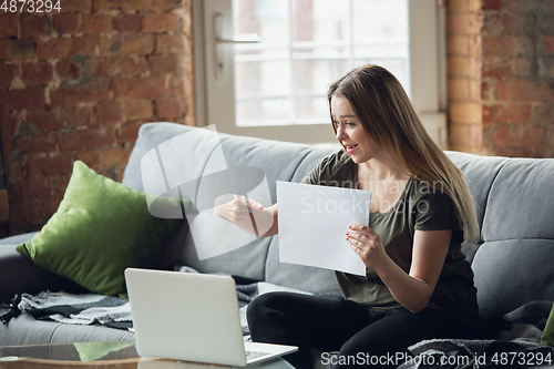 Image of Young woman, businesswoman working or studying at home, looking on computer screen, monitor, holding white sheet, whiteboard. Attented, concentrated. Copyspace. Top view.