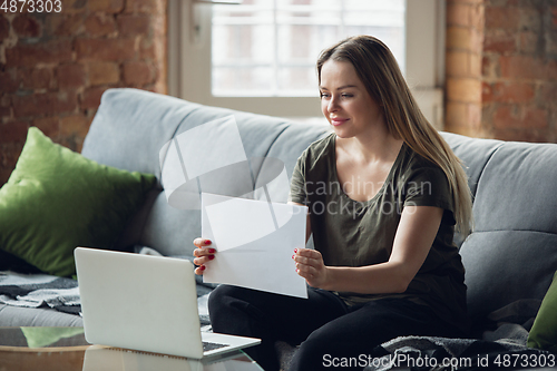 Image of Young woman, businesswoman working or studying at home, looking on computer screen, monitor, holding white sheet, whiteboard. Attented, concentrated. Copyspace. Top view.