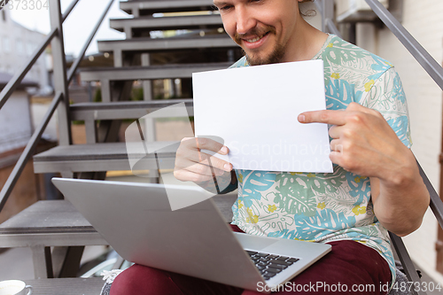 Image of Young man, businessman working outdoors, looking on computer screen, monitor, holding blank white sheet, whiteboard. Copyspace.