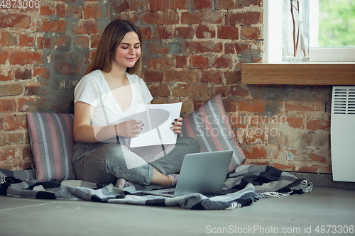 Image of Young woman, businesswoman working or studying at home, looking on computer screen, monitor, holding white sheet, whiteboard. Attented, concentrated. Copyspace. Top view.