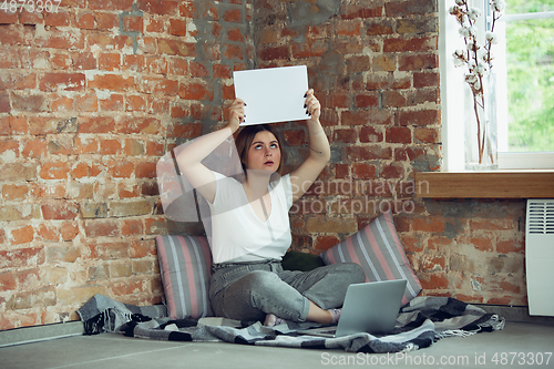 Image of Young woman, businesswoman working or studying at home, looking on computer screen, monitor, holding white sheet, whiteboard. Attented, concentrated. Copyspace. Top view.