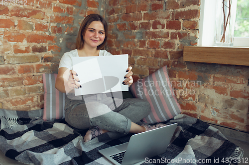 Image of Young woman, businesswoman working or studying at home, looking on computer screen, monitor, holding white sheet, whiteboard. Attented, concentrated. Copyspace. Top view.