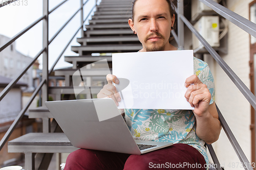 Image of Young man, businessman working outdoors, looking on computer screen, monitor, holding blank white sheet, whiteboard. Copyspace.