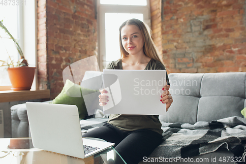 Image of Young woman, businesswoman working or studying at home, looking on computer screen, monitor, holding white sheet, whiteboard. Attented, concentrated. Copyspace. Top view.