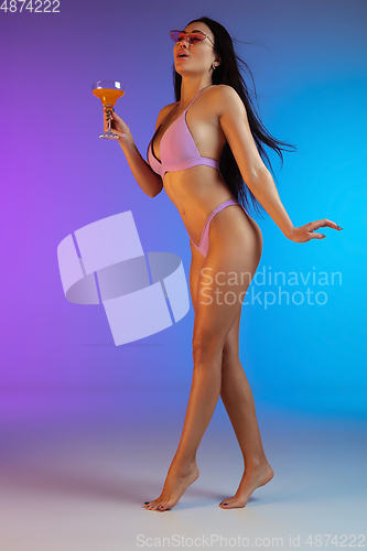 Image of Fashion portrait of young fit and sportive woman in purple luxury swimwear with cocktail and stylish sunglasses on gradient background. Perfect body ready for summertime.