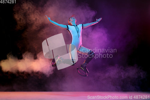 Image of Beautiful redhead woman in a white sportswear jumping in a kangoo jumps shoes isolated on gradient studio background in neon lighted smoke