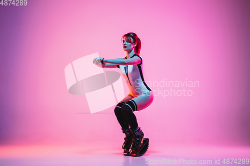 Image of Beautiful redhead woman in a white sportswear jumping in a kangoo jumps shoes isolated on gradient studio background in neon light